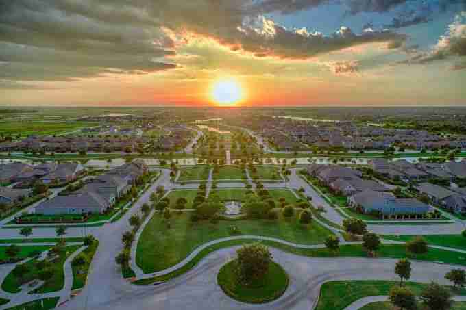 A vibrant aerial view of a Cypress neighborhood at sunset, featuring well-maintained homes, green spaces, and a dramatic sky with colorful clouds.