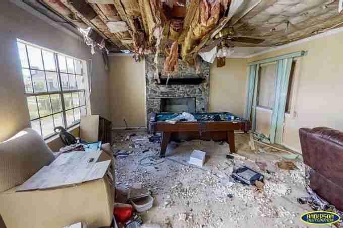 Photo of a severely water-damaged living room with a collapsed ceiling exposing wooden beams and insulation. Debris is scattered across the floor, with damaged furniture including a pool table, a chair, and a fireplace in the background. The text above reads 'How Can Water Damage Ruin Lives?' followed by a description of the structural, financial, and emotional impacts of water damage. The Anderson Restoration logo is displayed in the bottom right corner.