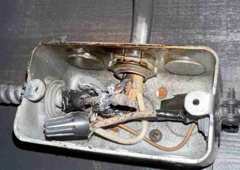 An open electrical junction box with severely damaged and burnt wiring inside. The wires appear melted and charred, with exposed copper and signs of overheating.
