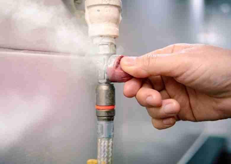 A close-up image of a person's hand turning a gas valve with steam or gas visibly leaking from the connection.