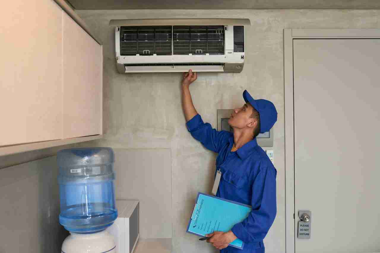 Technician in blue uniform inspecting a wall-mounted HVAC unit during a routine maintenance check inside a building.