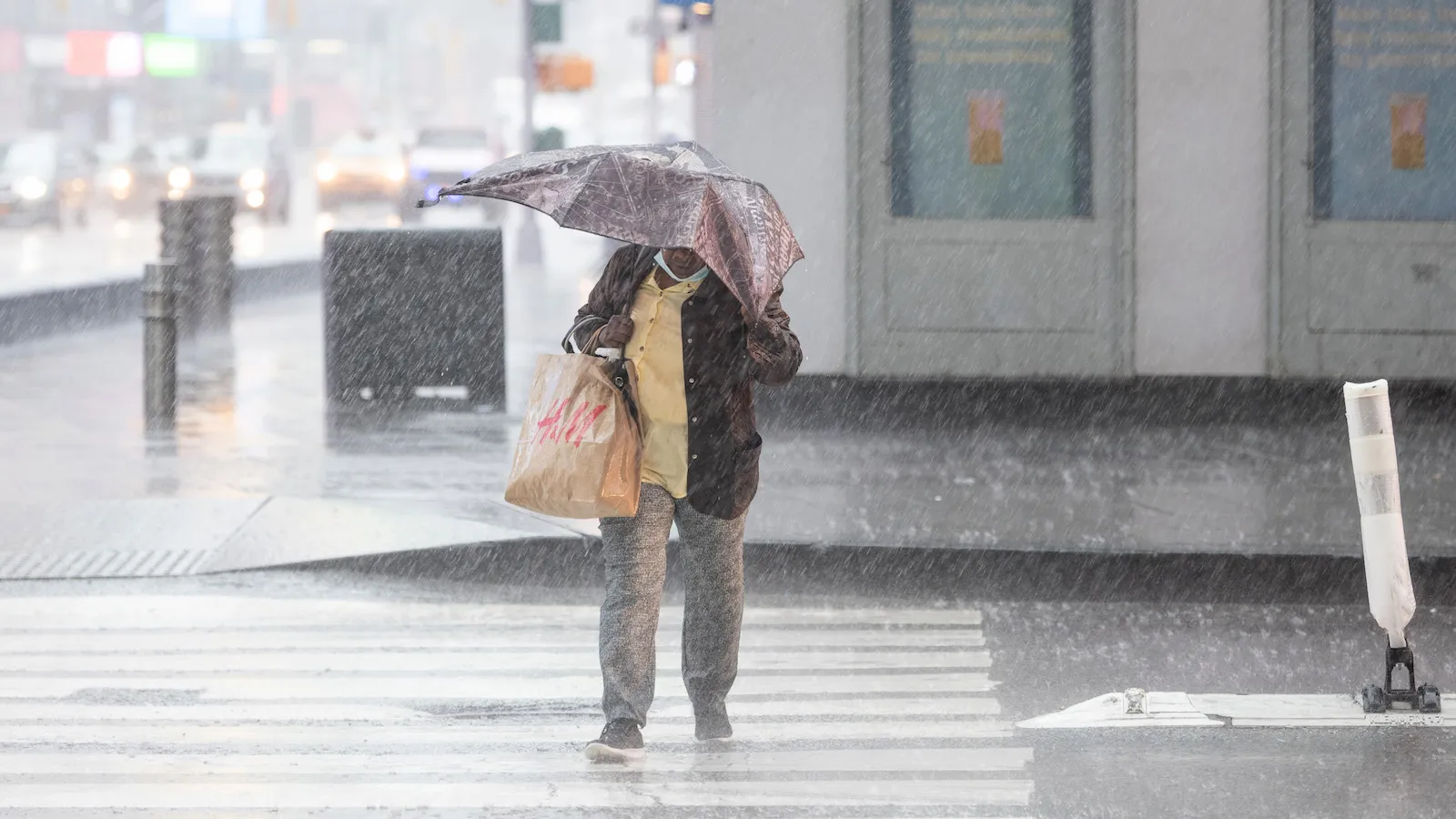A person crosses a city street in heavy rain, holding a damaged umbrella and a shopping bag, with blurred headlights and wet pavement in the background.