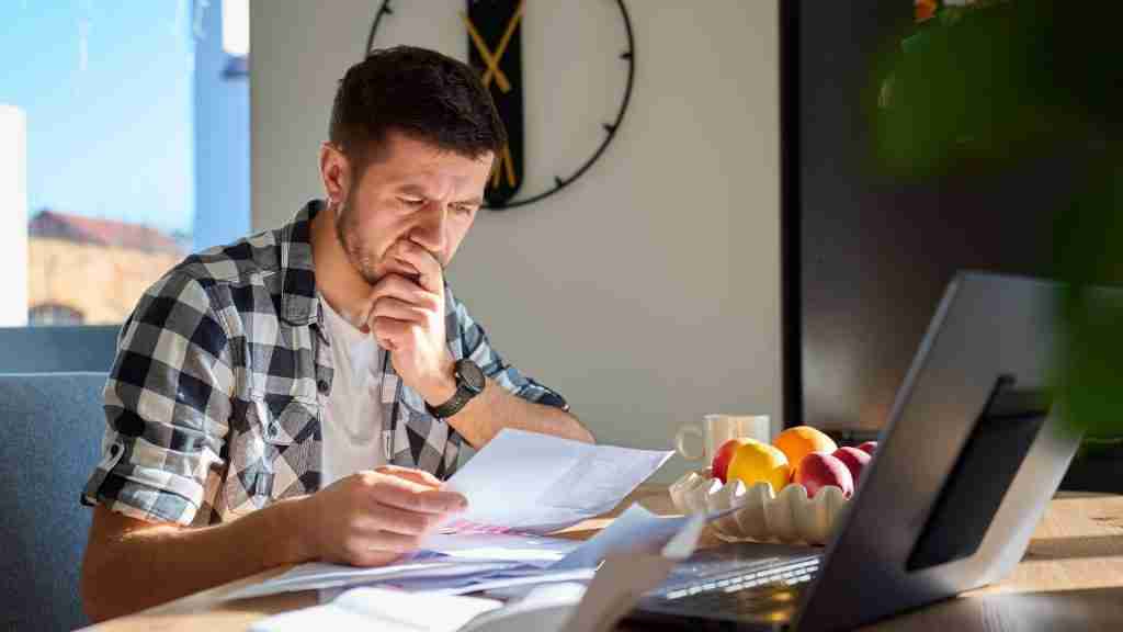 Homeowner at a kitchen table reviewing an insurance policy document with a laptop open showing a roof estimate