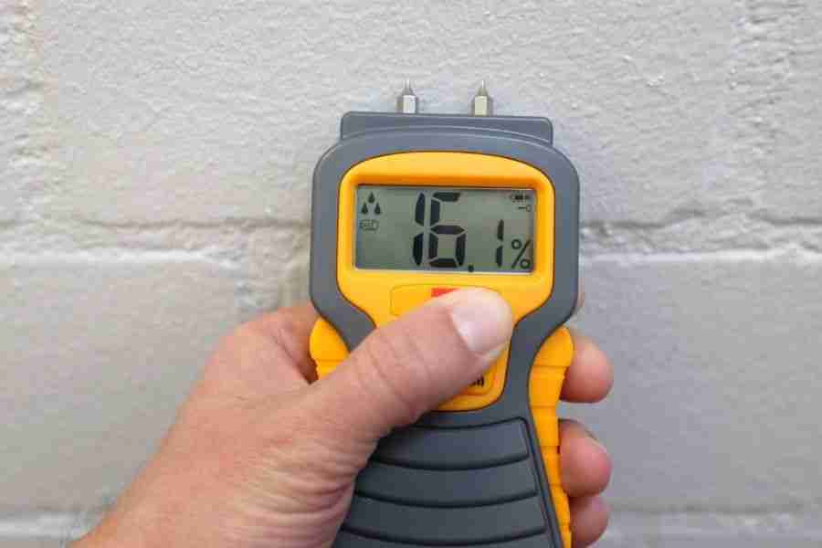 Yellow moisture meter pressed against a white painted brick wall reading 16.1 percent during water extraction.