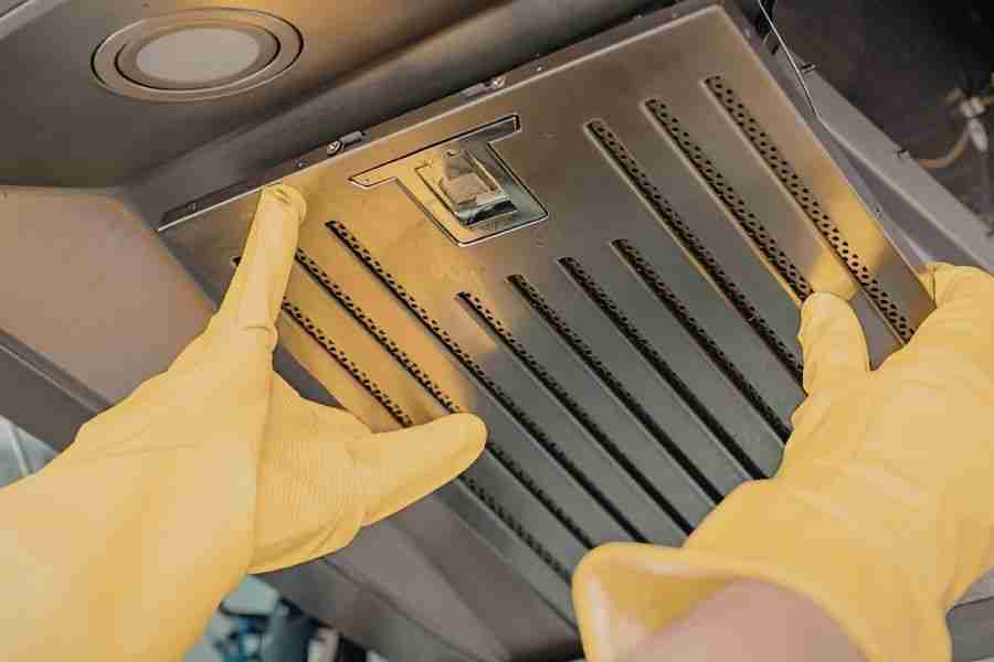 Gloved hands removing a stainless steel kitchen range hood filter during routine ventilation cleaning.