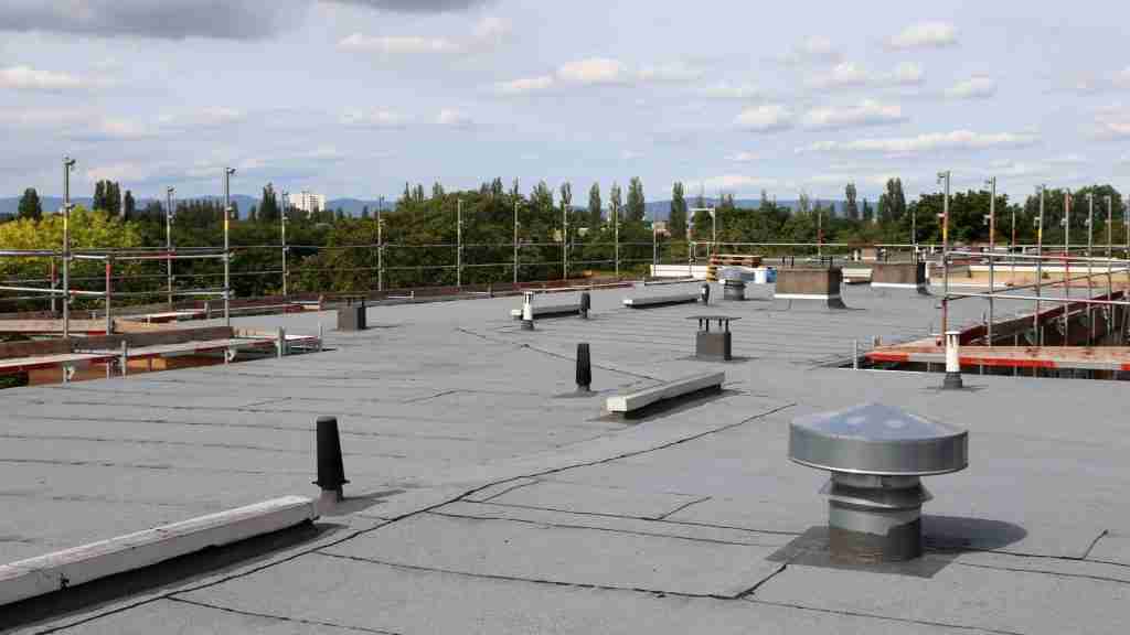 Roofing technician with clipboard and moisture meter performing a routine inspection on a large commercial flat roof, with drains, HVAC units, and parapet walls visible around him
